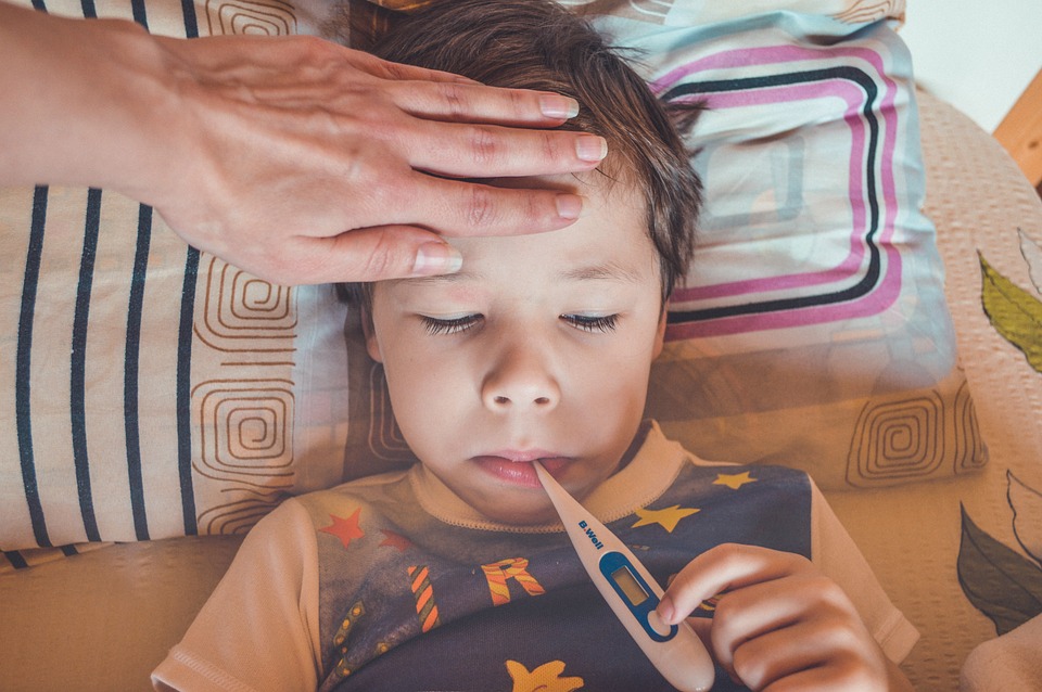 Bild på liten pojke som ligger i en säng med en febertermometer i munnen samtidigt spm en kvinna håller handen på hans panna